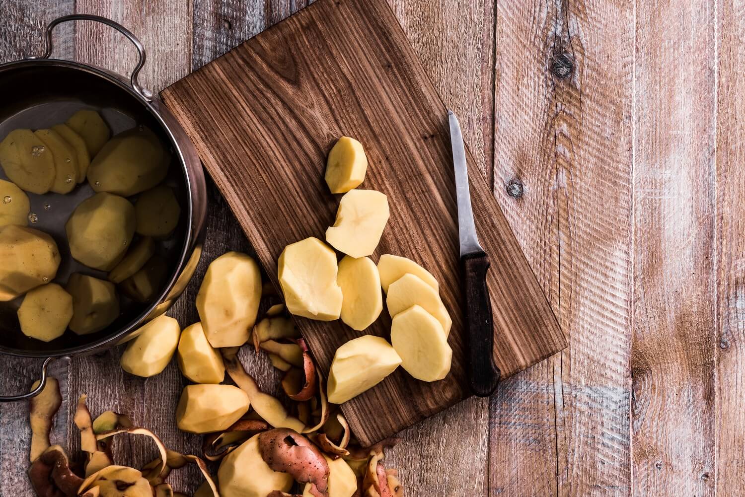 Kartoffeln werden auf einem Holzbrett geschält – daneben ein Topf mit gekochten Kartoffeln